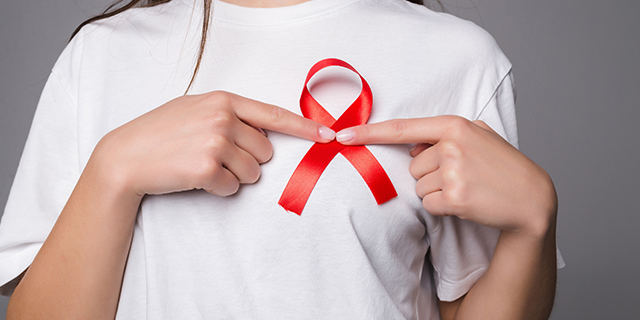 Mulher vestida de branco aponta para um laço vermelho em sua blusa, fazendo alusão ao Dezembro Vermelho